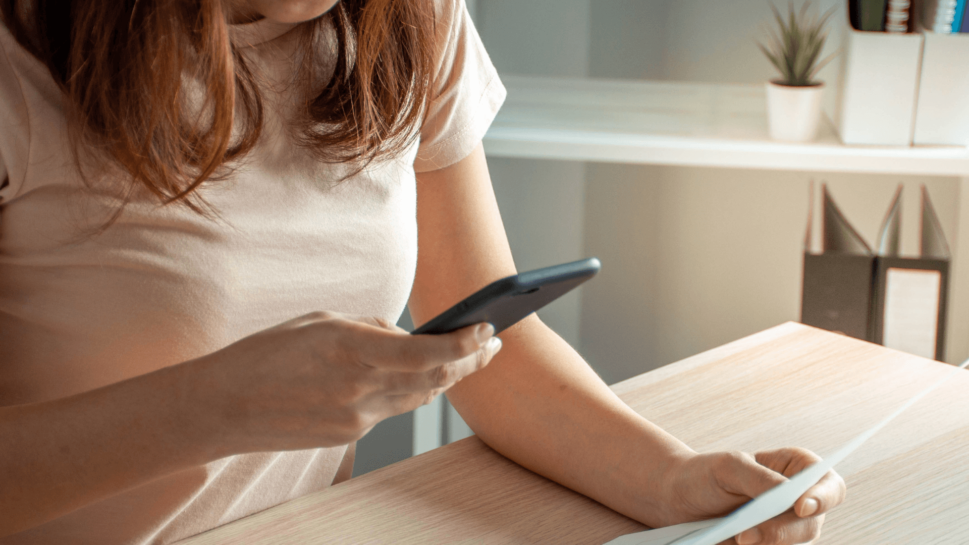 Mujer sentada a la mesa con el móvil en una mano y una factura de papel en la otra, revisando el gasto de su línea de teléfono.
