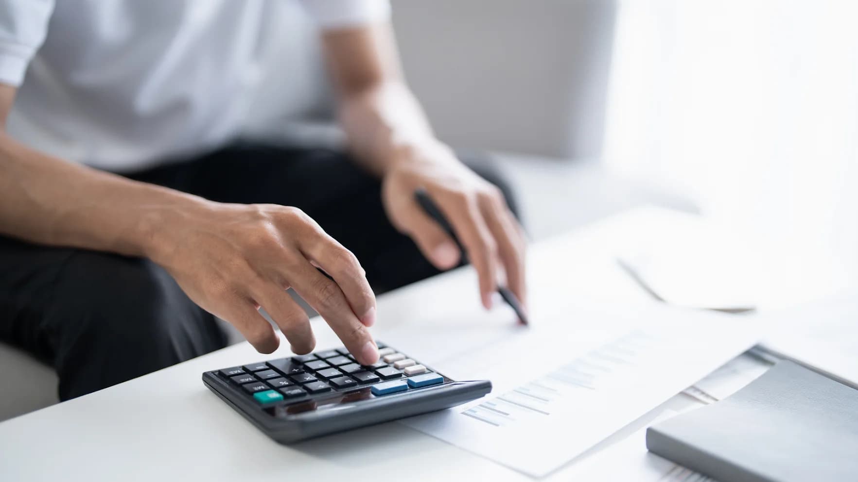 Persona usando una calculadora sobre una mesa blanca junto a un documento con gráficas y un bolígrafo.