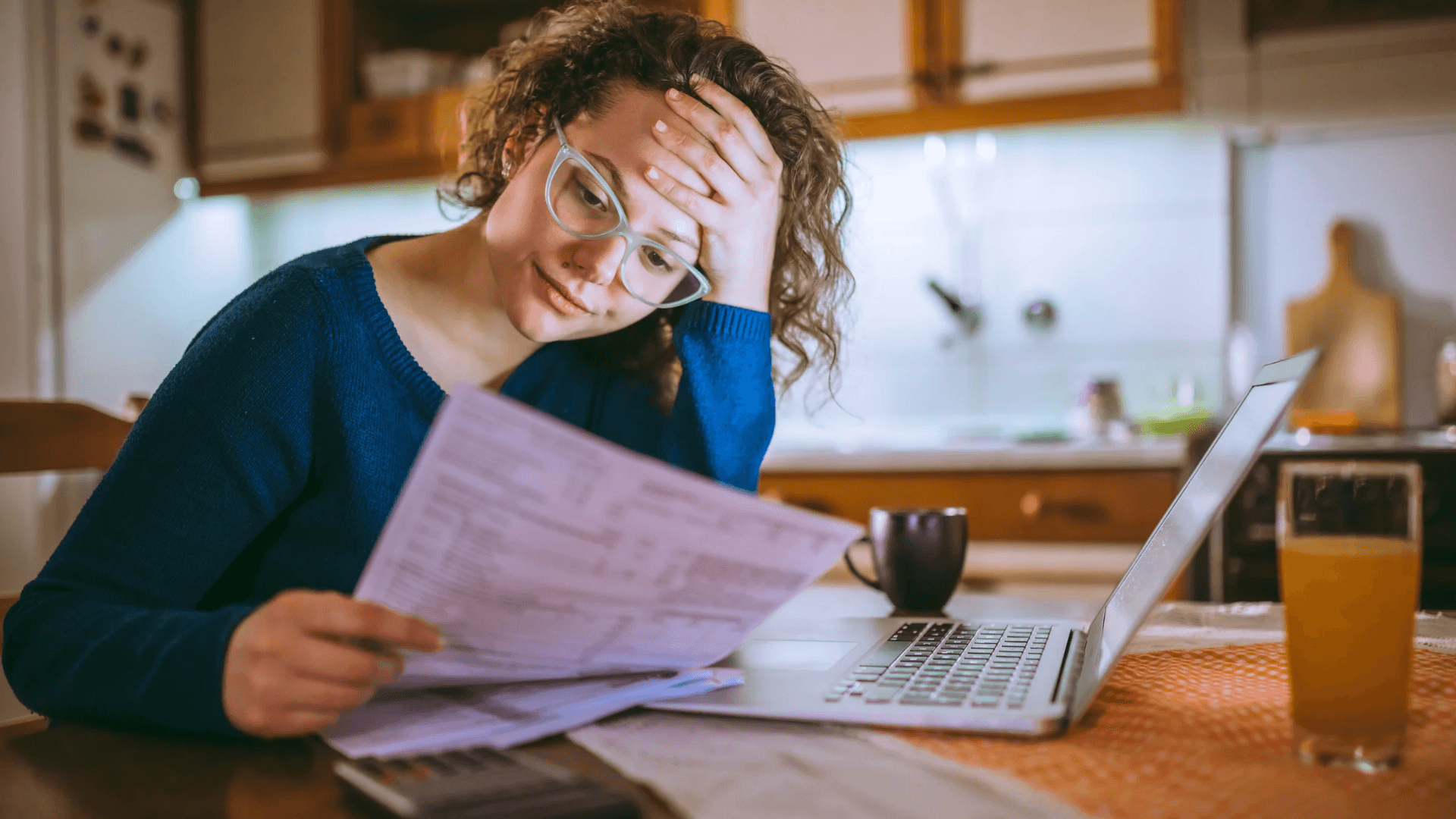 Mujer con gafas revisando con preocupación una factura de la luz en la cocina, junto a un portátil y una calculadora.